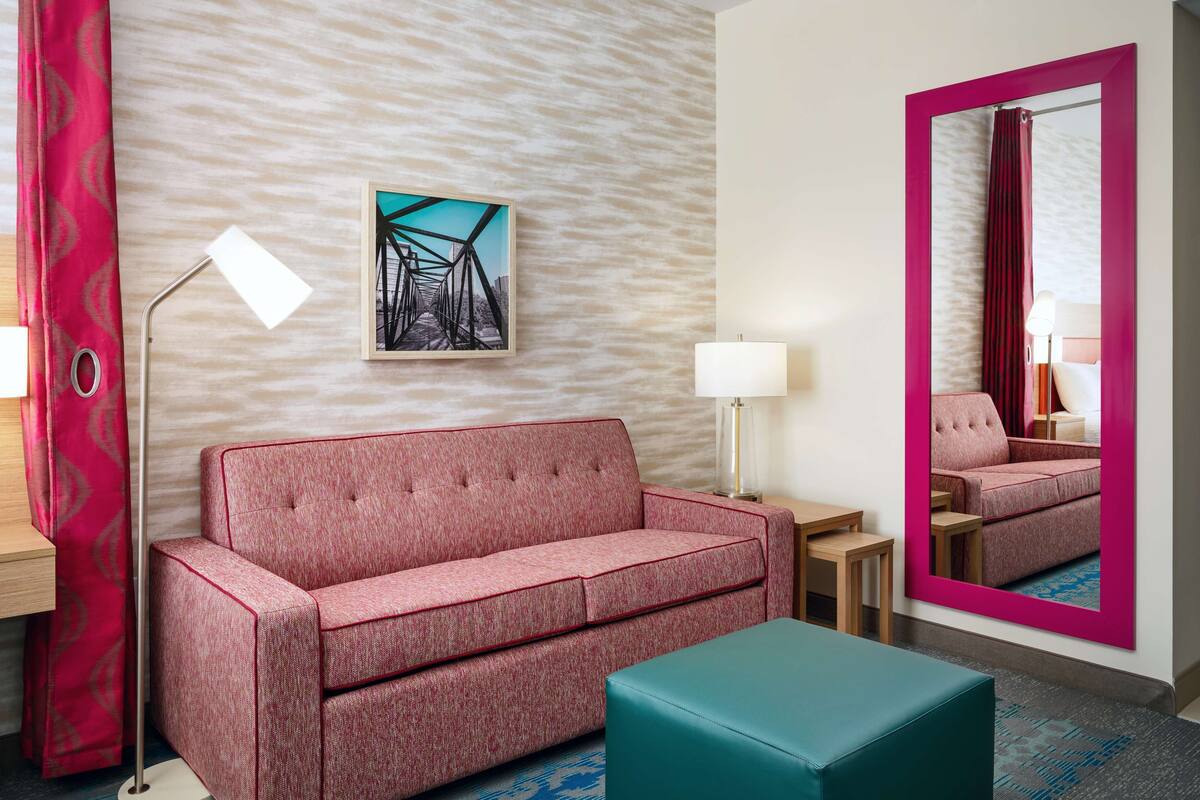 Une chambre d’hôtel avec un canapé rouge, un pouf turquoise, une lampe sur pied, une table et un tableau au mur.