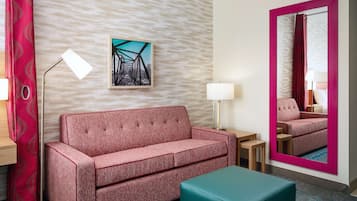 Quarto de hotel com um sofá vermelho, um banquinho verde-água, uma luminária de chão, uma mesa e um quadro na parede.
