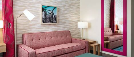 A hotel room with a red sofa, a teal ottoman, a floor lamp, a table, and a painting on the wall.