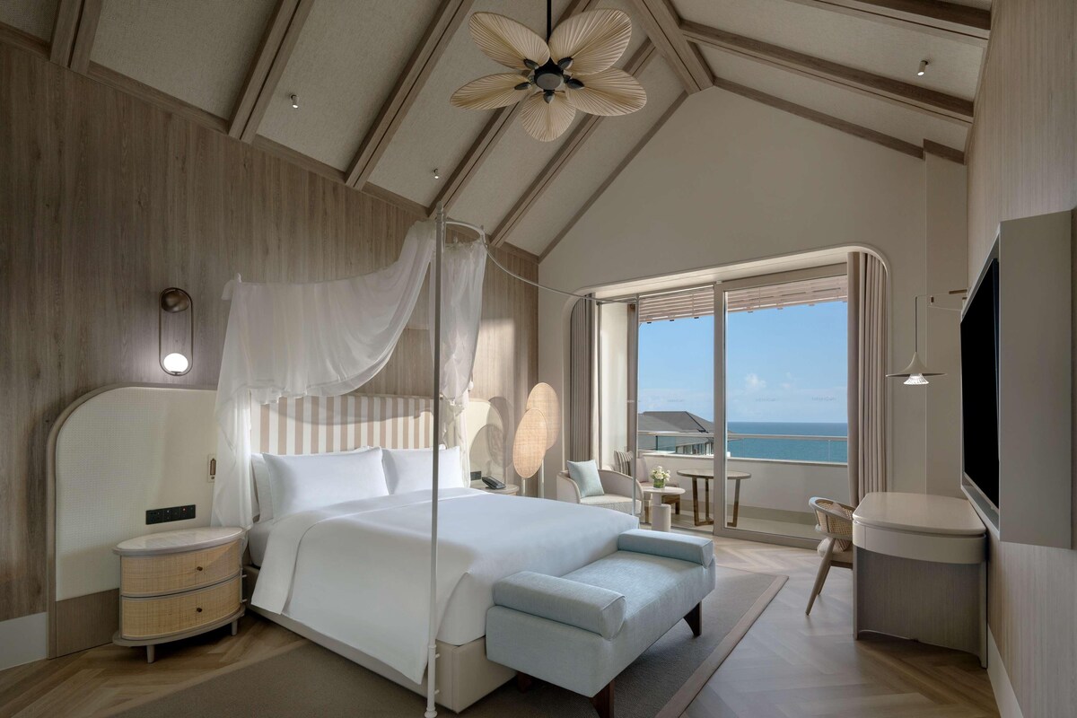 A bedroom with a large bed, a bedside table, a nightstand, a TV, and a balcony with a view of the sea.