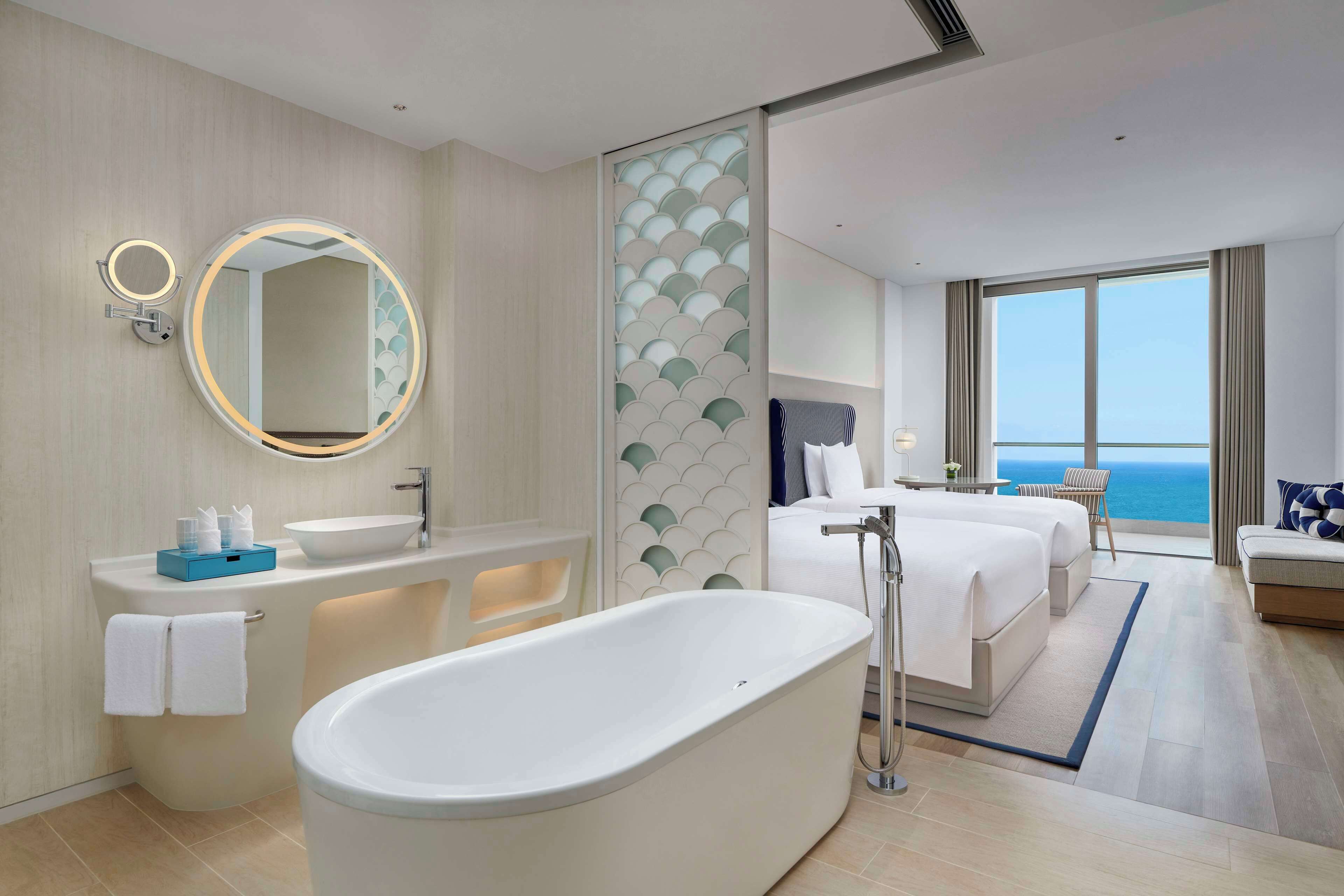 Una habitación de hotel moderna con una bañera, un espejo redondo grande, un tocador con lavamanos y una cama con vista al océano.