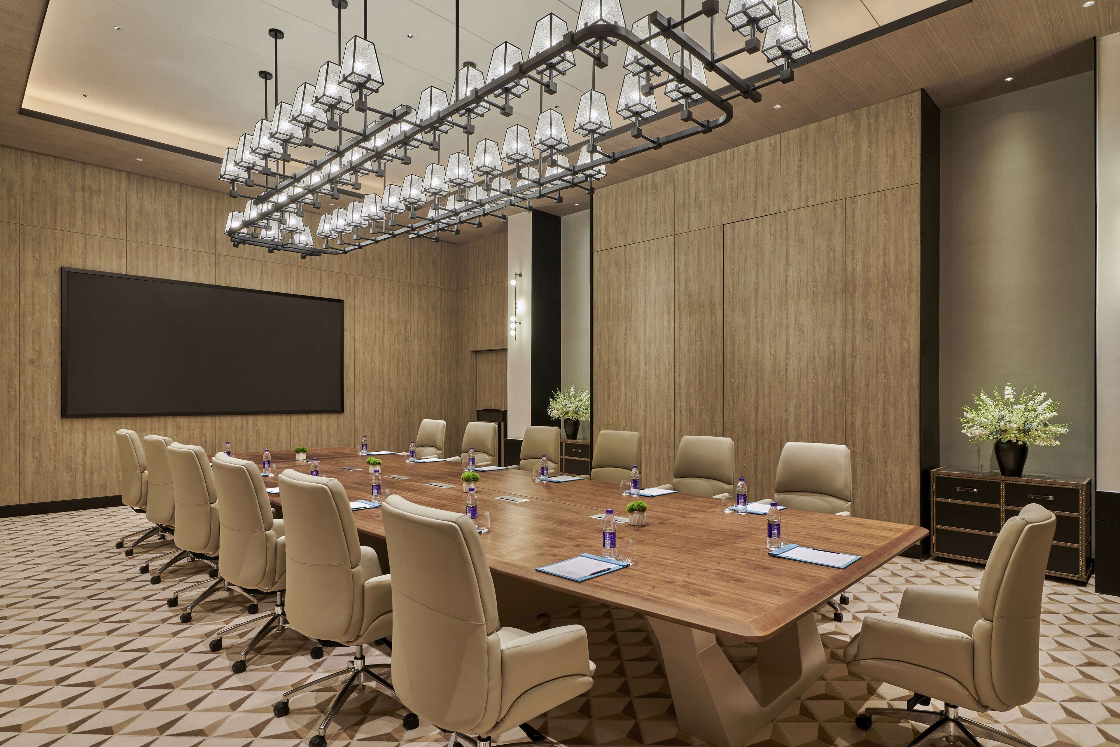 A conference room with a large wooden table, beige chairs, a projector screen, and a modern chandelier.