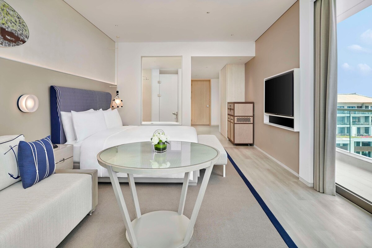 A hotel room with a bed, a round glass table, a TV, and a view of the outside.
