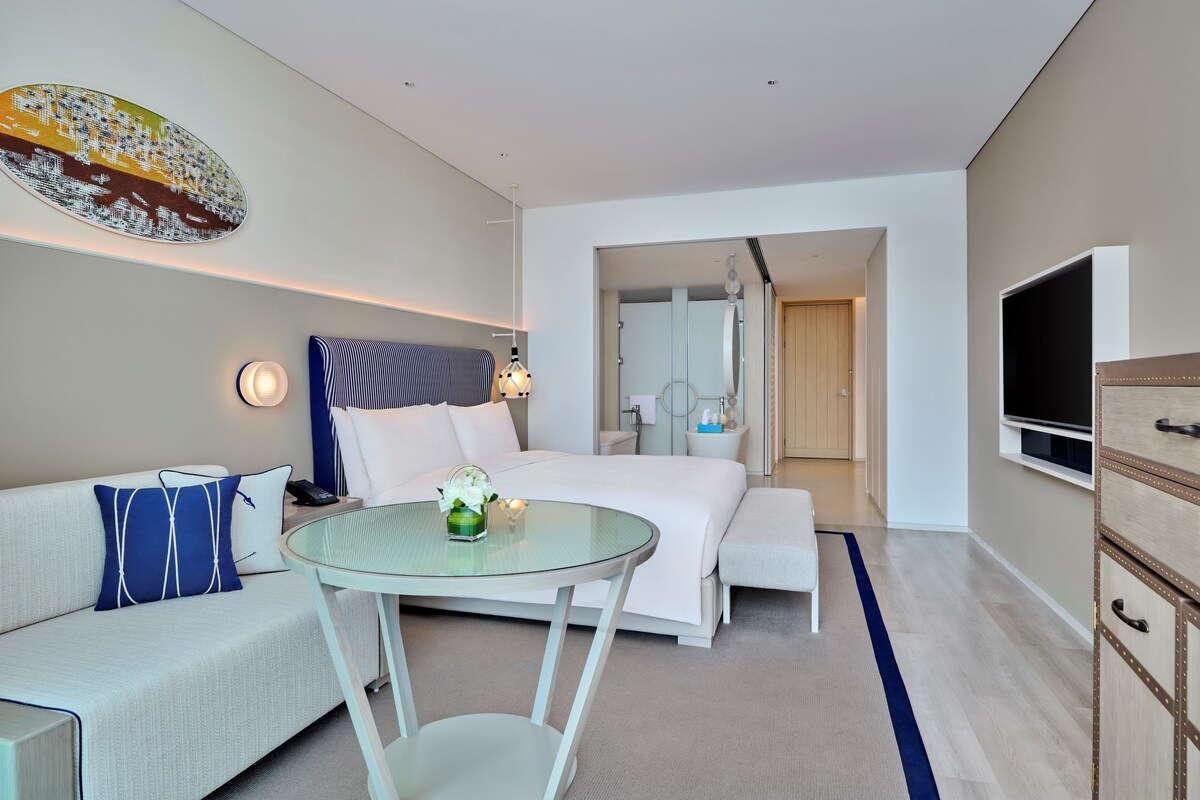 A modern hotel room with a bed, a glass table, a sofa, a TV, and a large artwork on the wall.