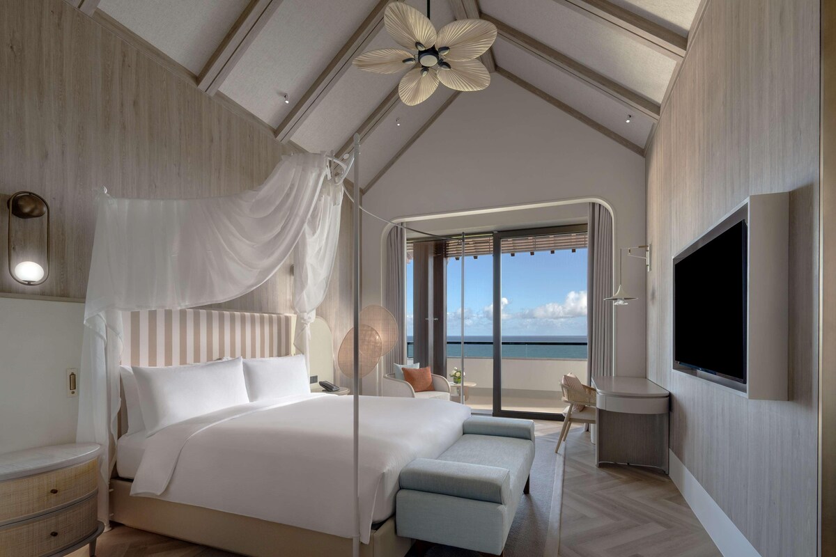 A modern bedroom with a large bed, a TV, a ceiling fan, and a view of the ocean.