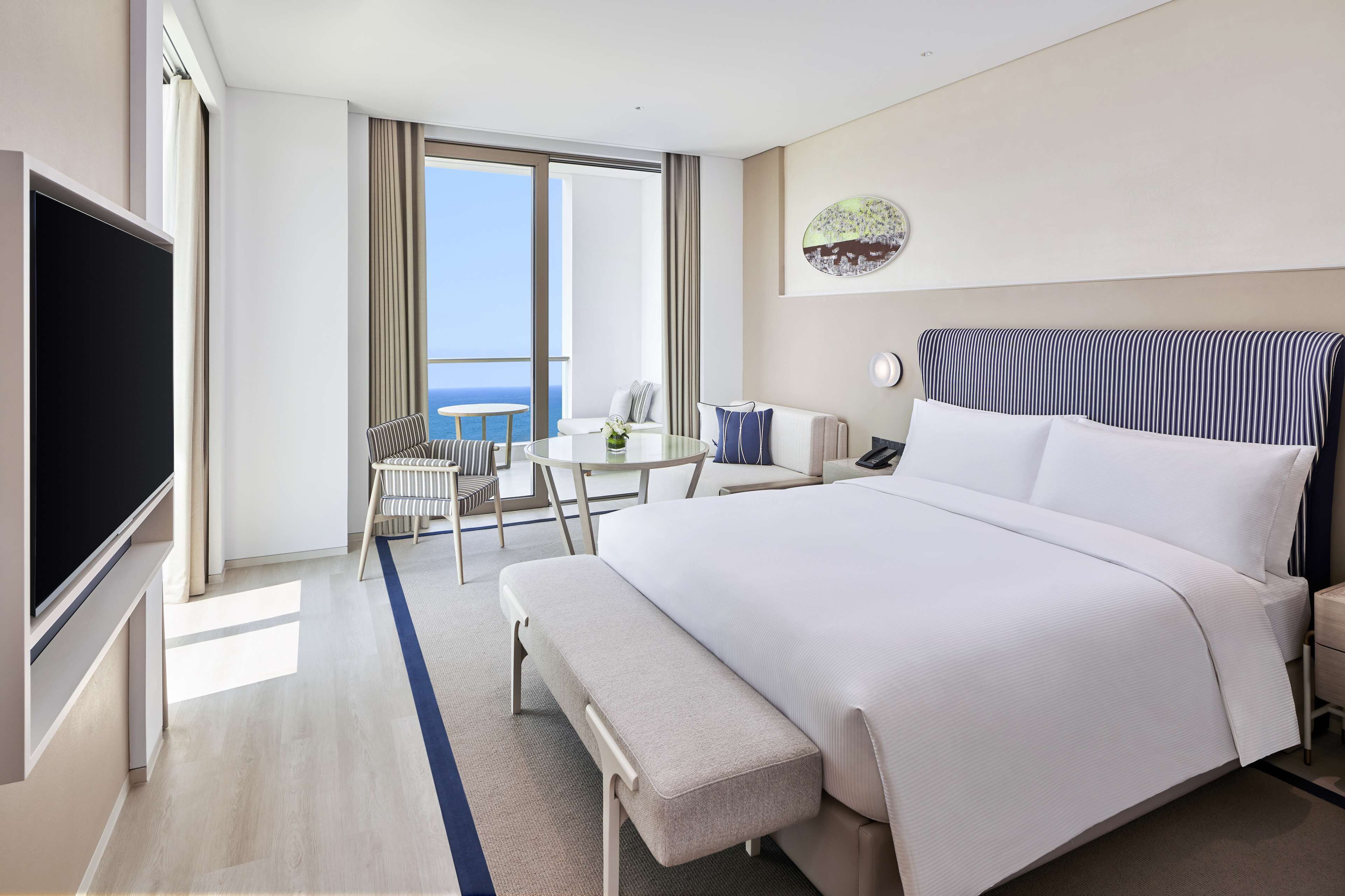 A hotel room with a large bed, a desk with a chair, a TV, and a view of the ocean.