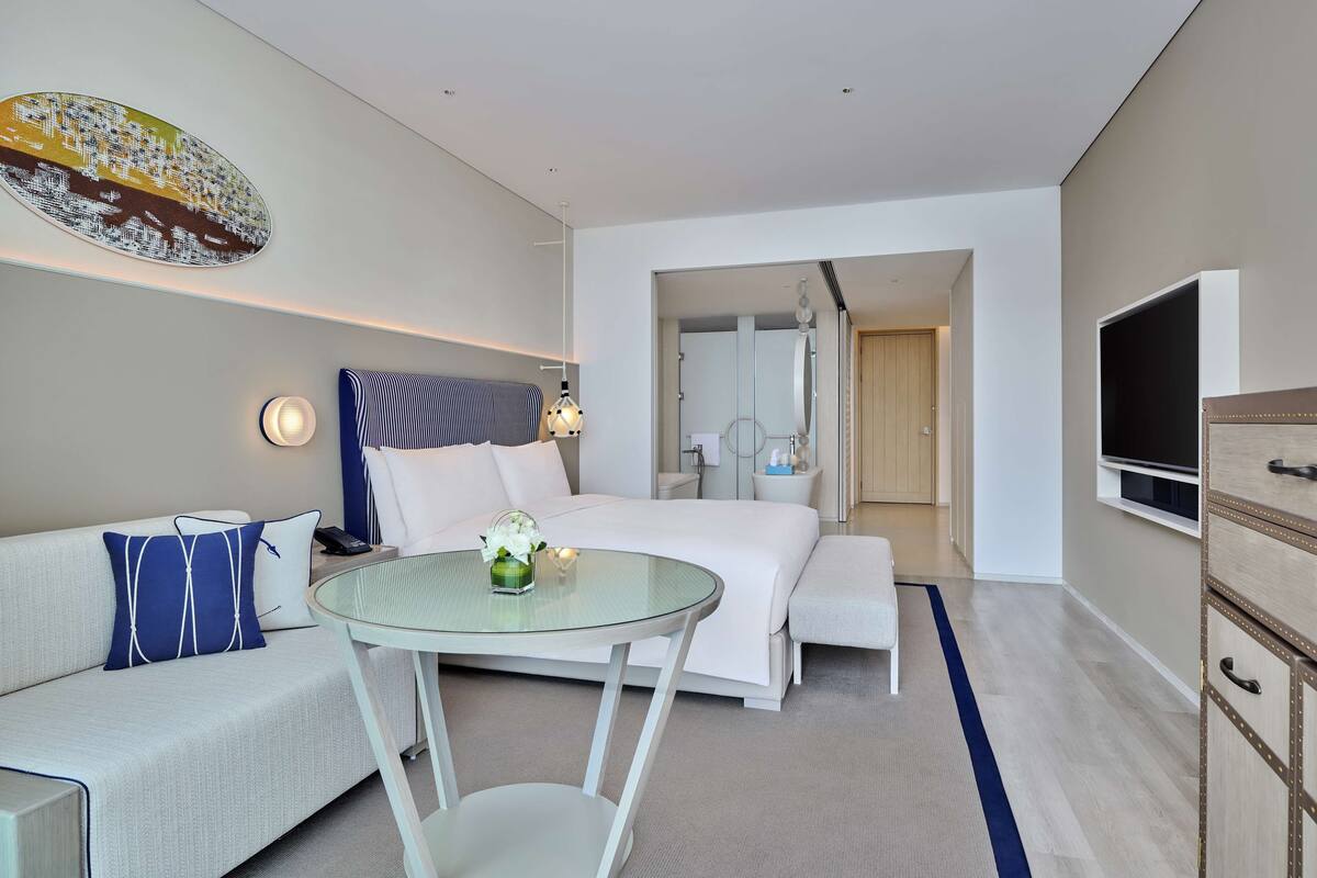 Una habitación de hotel moderna con una cama, una mesa de vidrio, un sofá, un televisor y un baño visible a través de una puerta abierta.