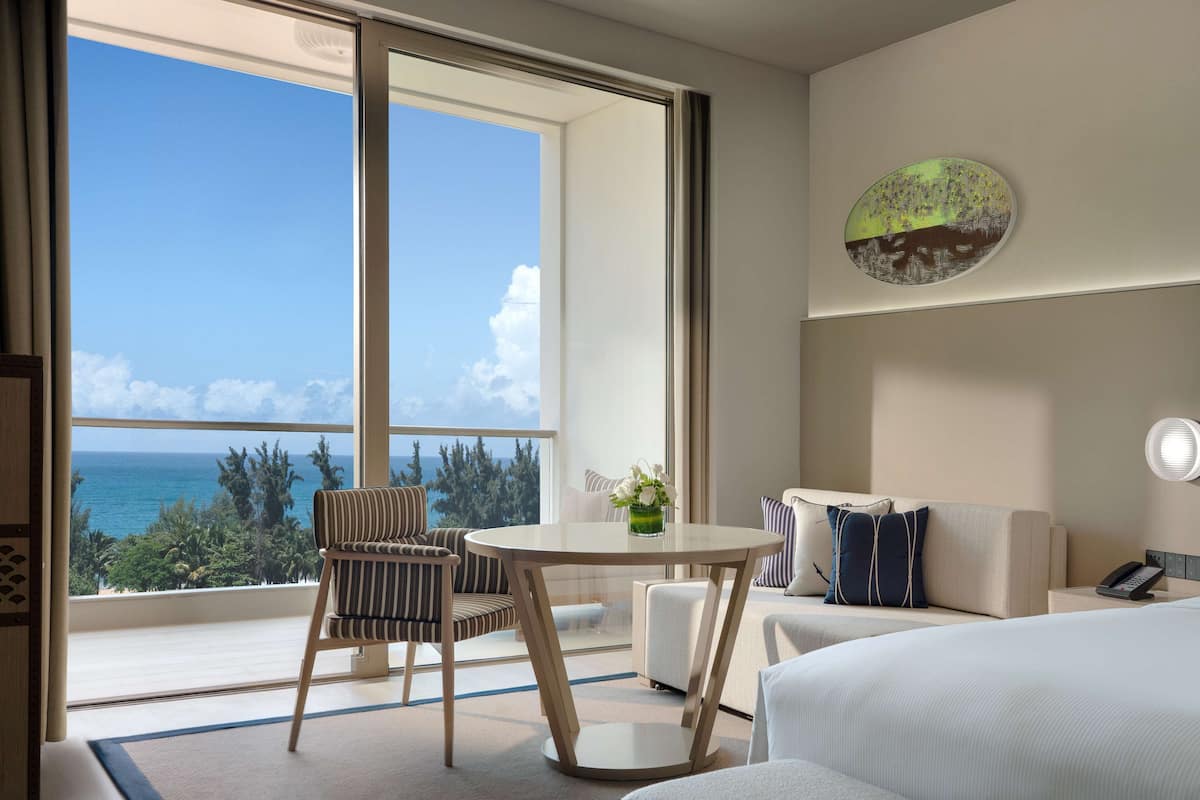 Habitación de hotel con un ventanal amplio que ofrece vistas al océano y árboles, una mesa redonda con un jarrón de flores, un sofá y una cama.
