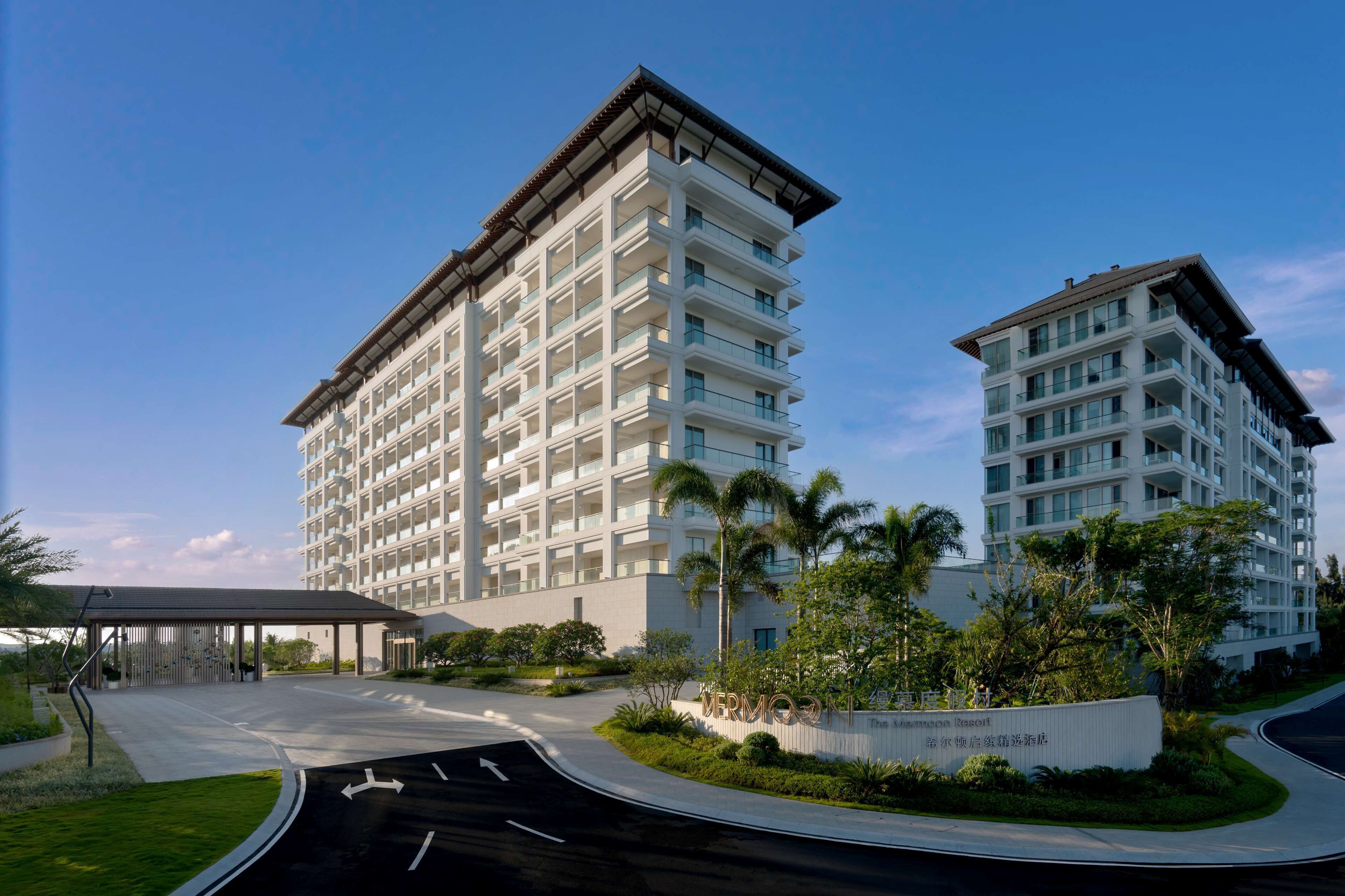 Um hotel moderno com vários andares, entrada coberta e área ajardinada com palmeiras.