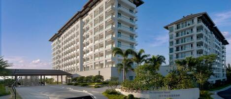 Um hotel moderno com vários andares, entrada coberta e área ajardinada com palmeiras.