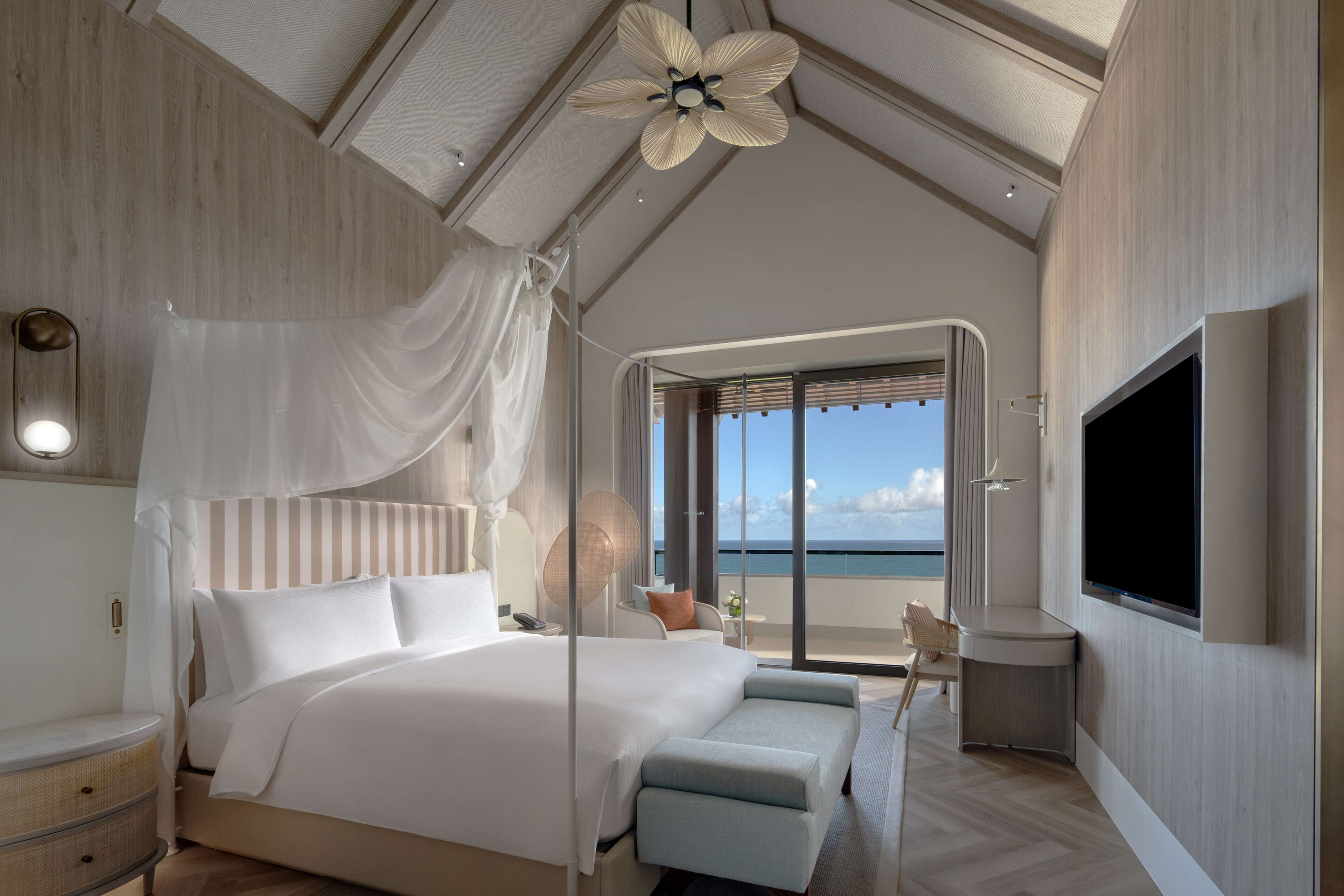 A modern bedroom with a large bed, a ceiling fan, a TV, and a balcony with a view of the sea.