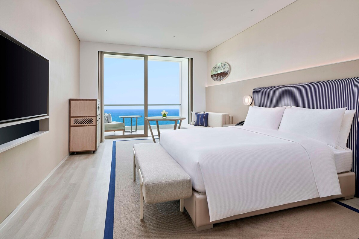 Habitación de hotel con una cama grande, televisión de pantalla plana, escritorio y vistas al océano a través de amplias ventanas.