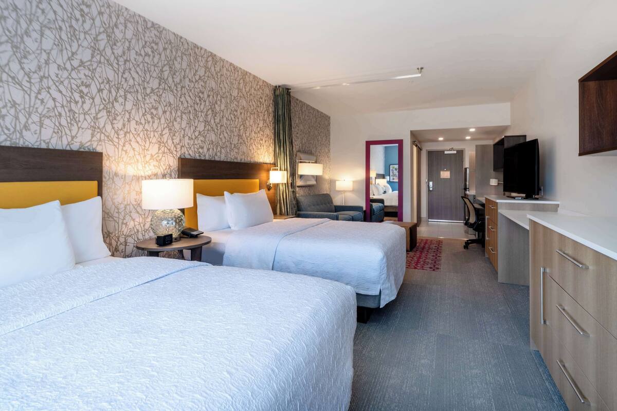 A hotel room with two beds, a desk, a TV, and a bathroom visible through a door.
