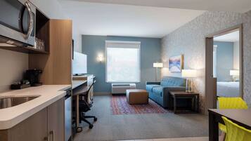A compact hotel room with a kitchenette, a sofa, a small table, and a window with a view.