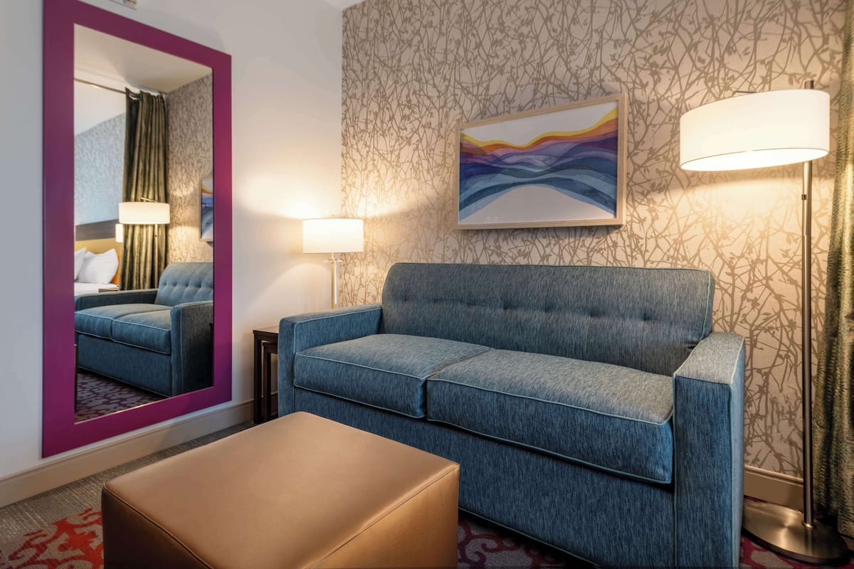 A hotel room with a blue sofa, a matching ottoman, a large mirror, a painting, and two floor lamps.