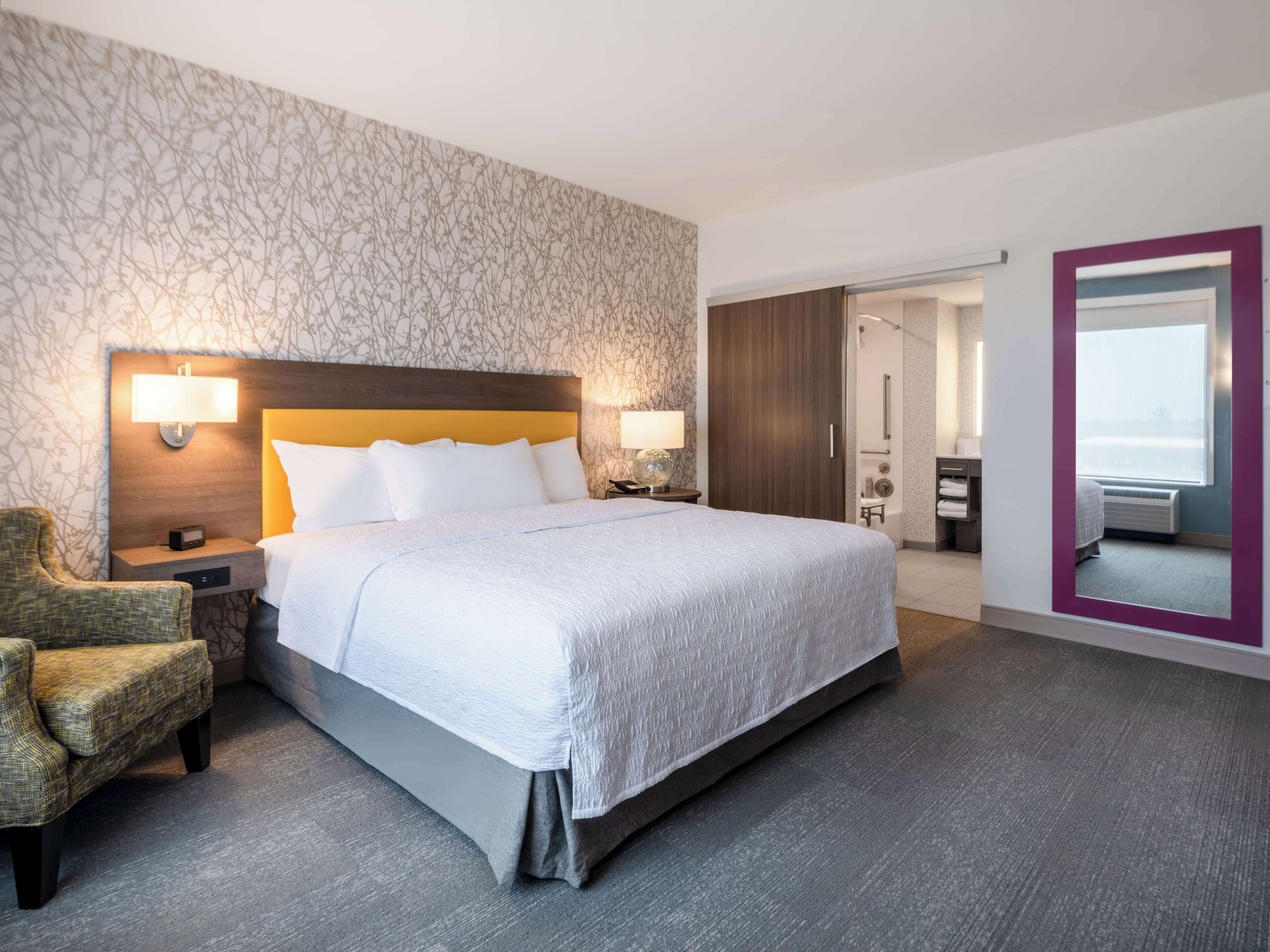 A hotel room with a large bed, a nightstand, a chair, a bathroom with a shower, and a view of the outside.