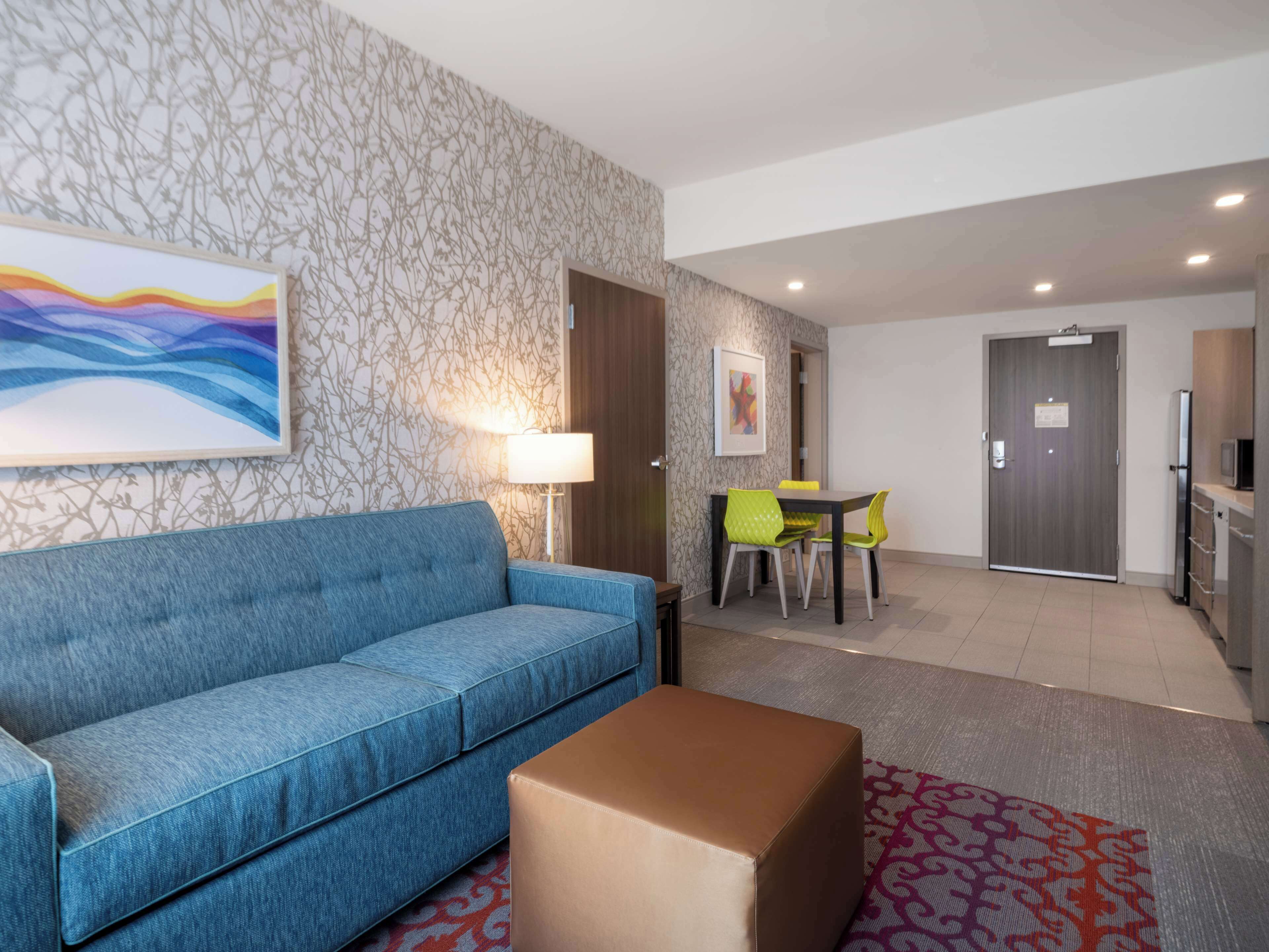 A hotel room with a blue sofa, a dining table with yellow chairs, a wooden cabinet, and a television.