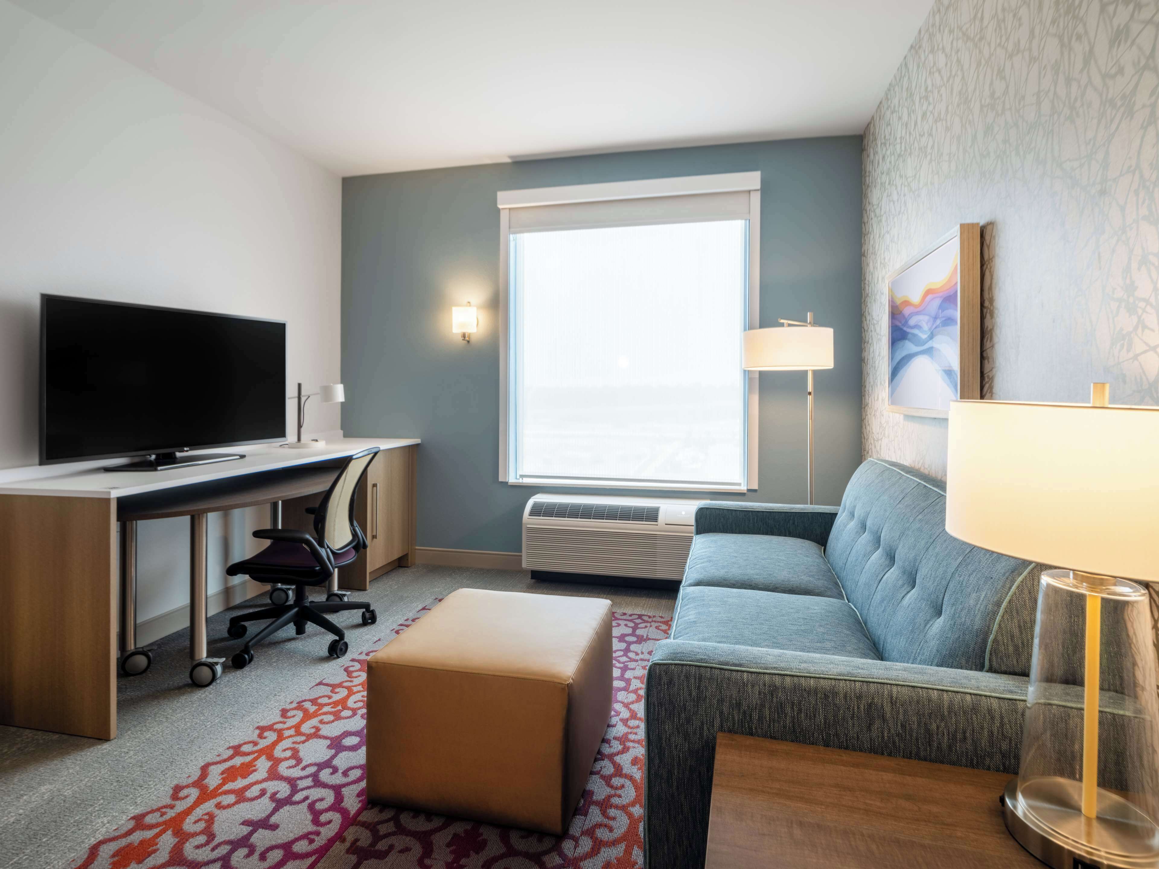 Quarto de hotel com televisão de tela plana, escrivaninha com cadeira, sofá, luminária de chão e uma janela grande.