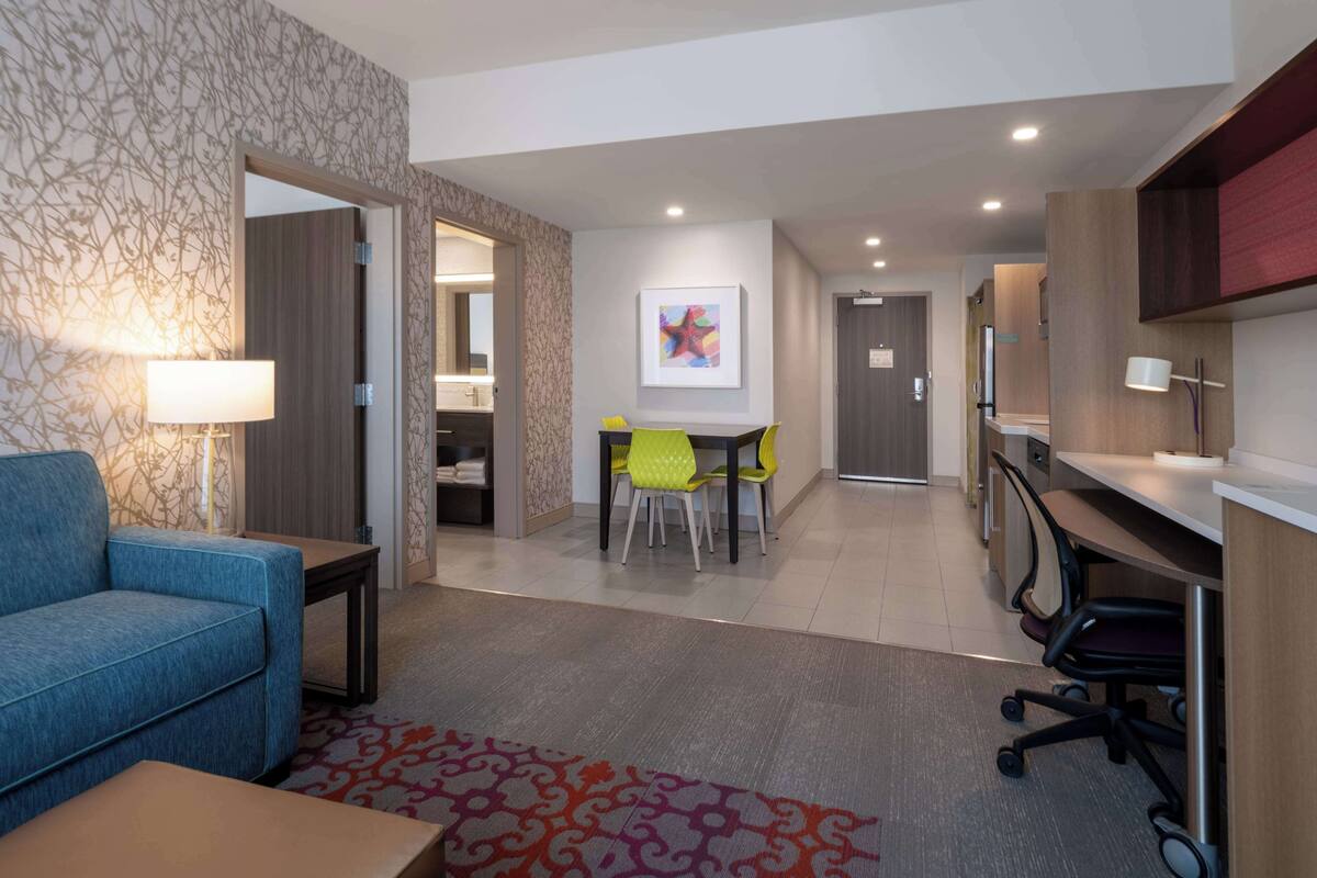 A hotel room with a blue sofa, a small table, a desk with a chair, a bathroom with a mirror, and a dining area with a table and chairs.