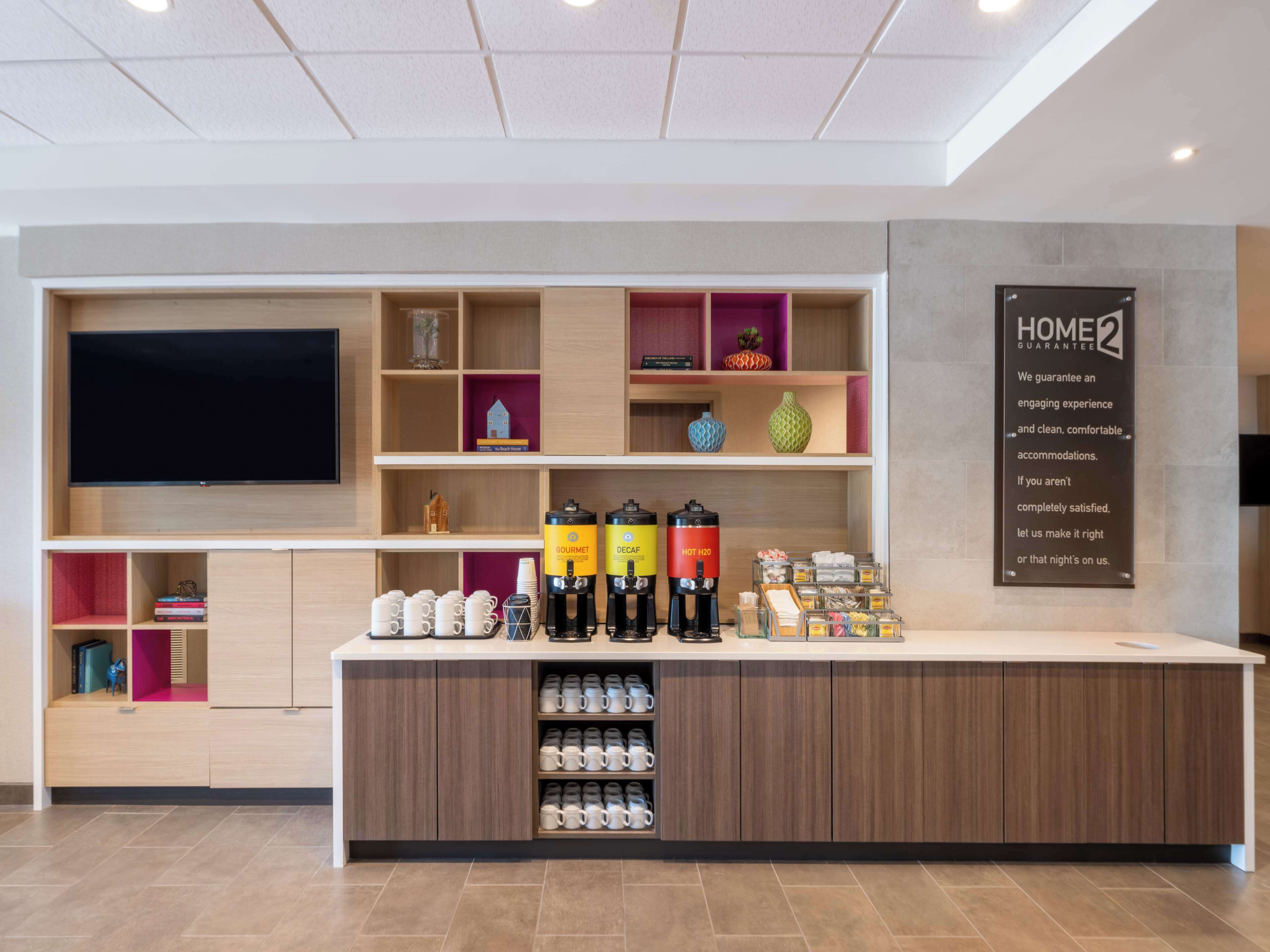Una moderna area reception hotel con una TV a schermo piatto, un angolo caffè e un cartello con la scritta 'Home2 Guarantee'.