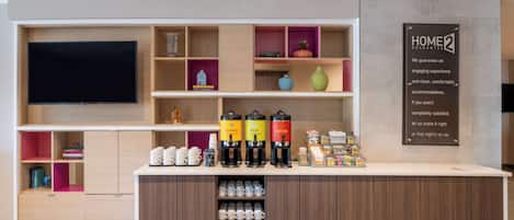 Una moderna area reception hotel con una TV a schermo piatto, un angolo caffè e un cartello con la scritta 'Home2 Guarantee'.