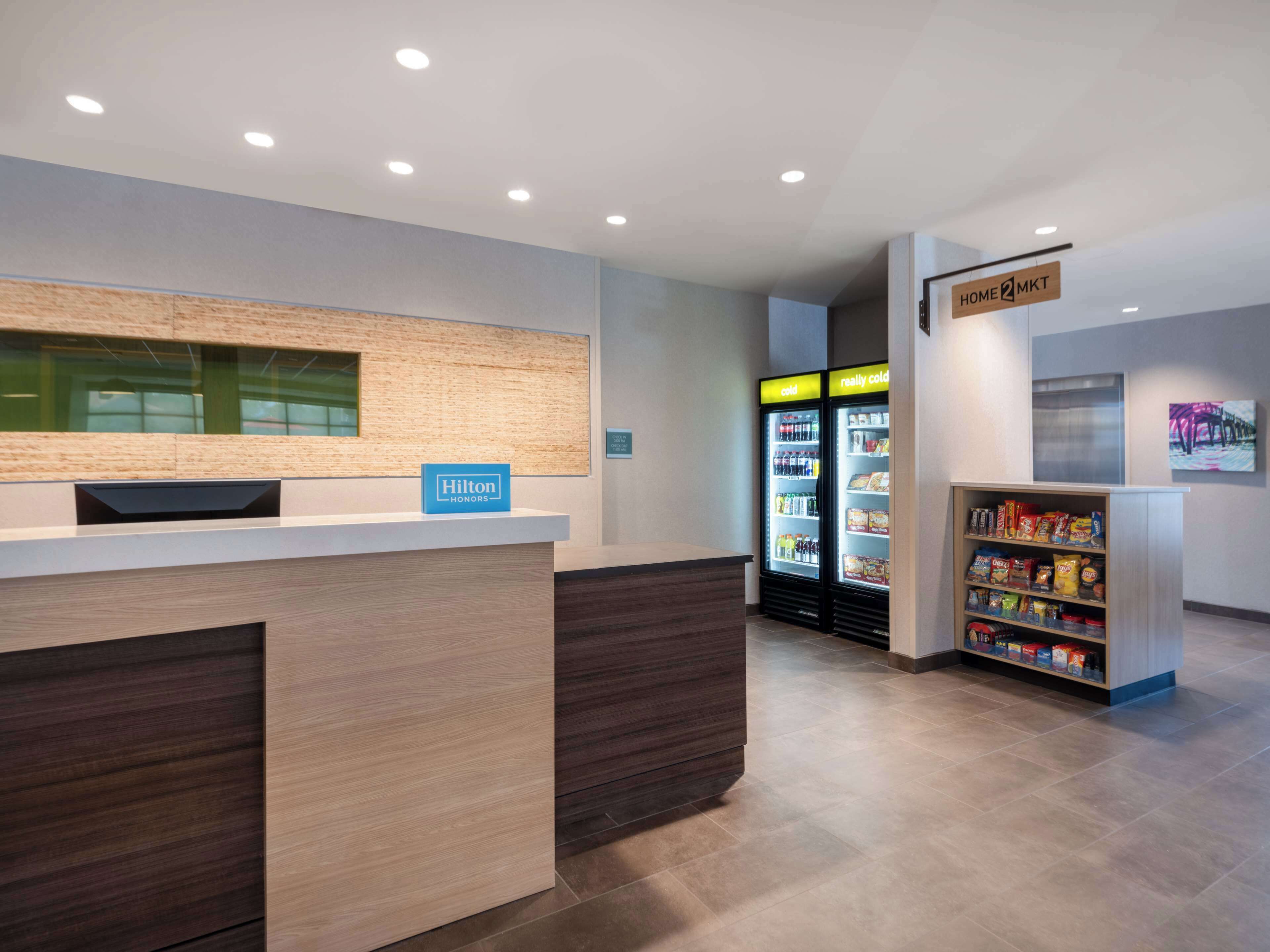 A hotel reception area with a wooden reception desk, a minibar, a cold beverage refrigerator, and a snack shelf.