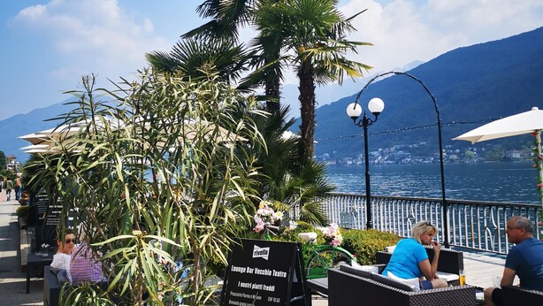 An outdoor seating area with a signboard, palm trees, and a view of the water. - Ca Sistina Blue (Morcote)