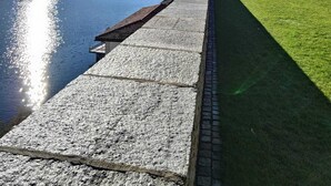 A waterfront promenade with a stone pathway, grassy areas, and a building with a red roof. - Ca Sistina Blue (Morcote)