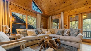 A cozy cabin interior with a sectional sofa, wooden coffee table, and large windows offering a view of the forest.