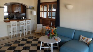 Un salon avec un canapé bleu, une petite table ronde ornée d’un vase de fleurs, un coin cuisine avec un comptoir-bar et des chaises blanches, et un sol à carreaux.