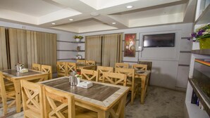 Uma sala de jantar com mesas e cadeiras de madeira, uma TV na parede e plantas decorativas.