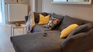 Ein modernes Wohnzimmer mit einer grauen Eckcouch, gelben Kissen, einem kleinen runden Tisch mit Pflanze und einer großen Wanduhr.