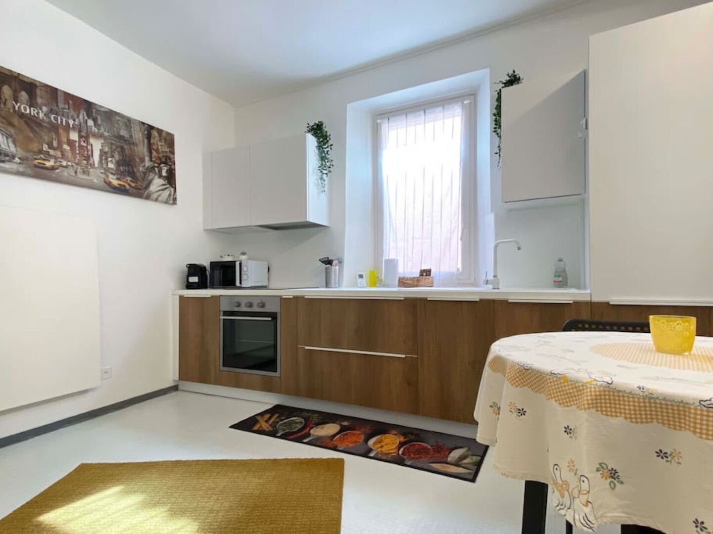 A compact kitchen with a microwave, oven, and sink. There's a window with sheer curtains, a small plant, and a yellow bowl on the table.