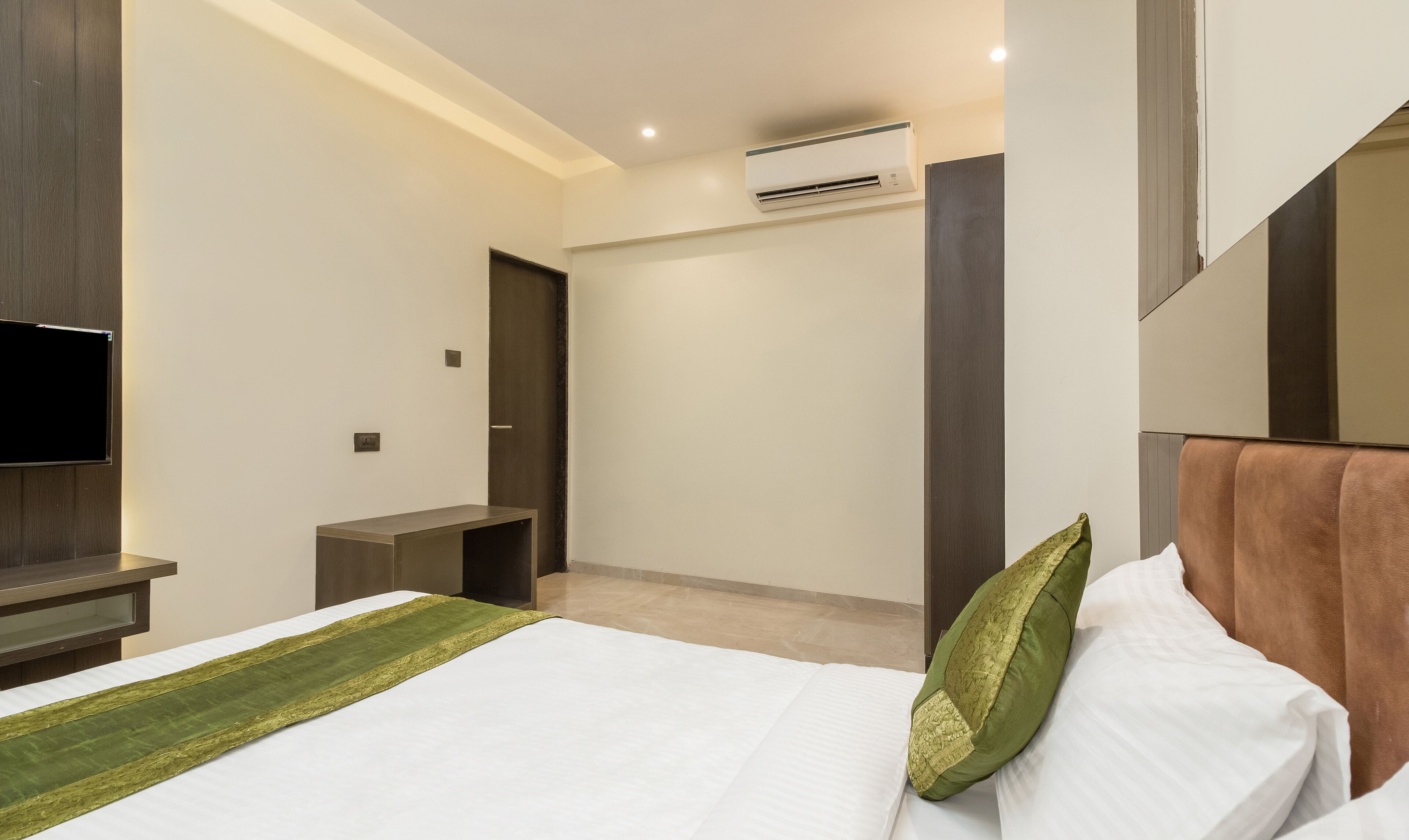 A modern hotel room with a bed, a flat-screen TV mounted on the wall, a small seating area, and an air conditioning unit.