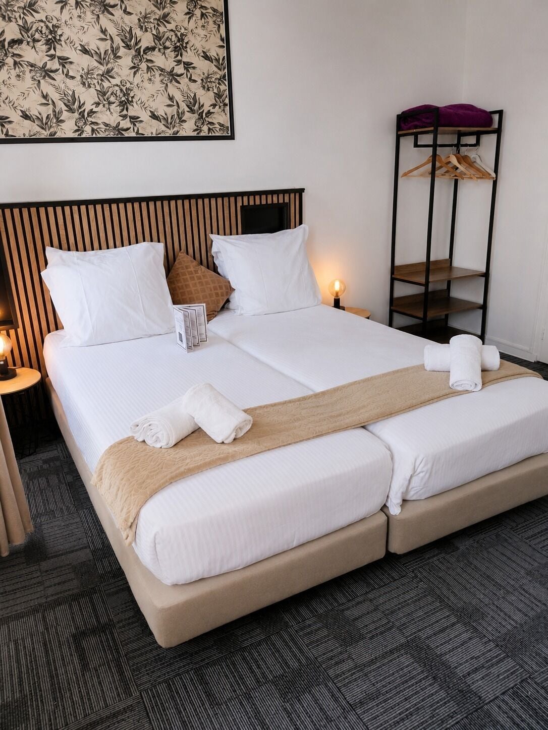 Habitación de hotel con una cama grande, dos mesitas de noche, un estante con perchas en la pared y una obra de arte floral enmarco.