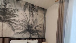 Una habitación de hotel con cama, mesita de noche, lámpara, un cuadro de palmeras y una ventana con cortinas.