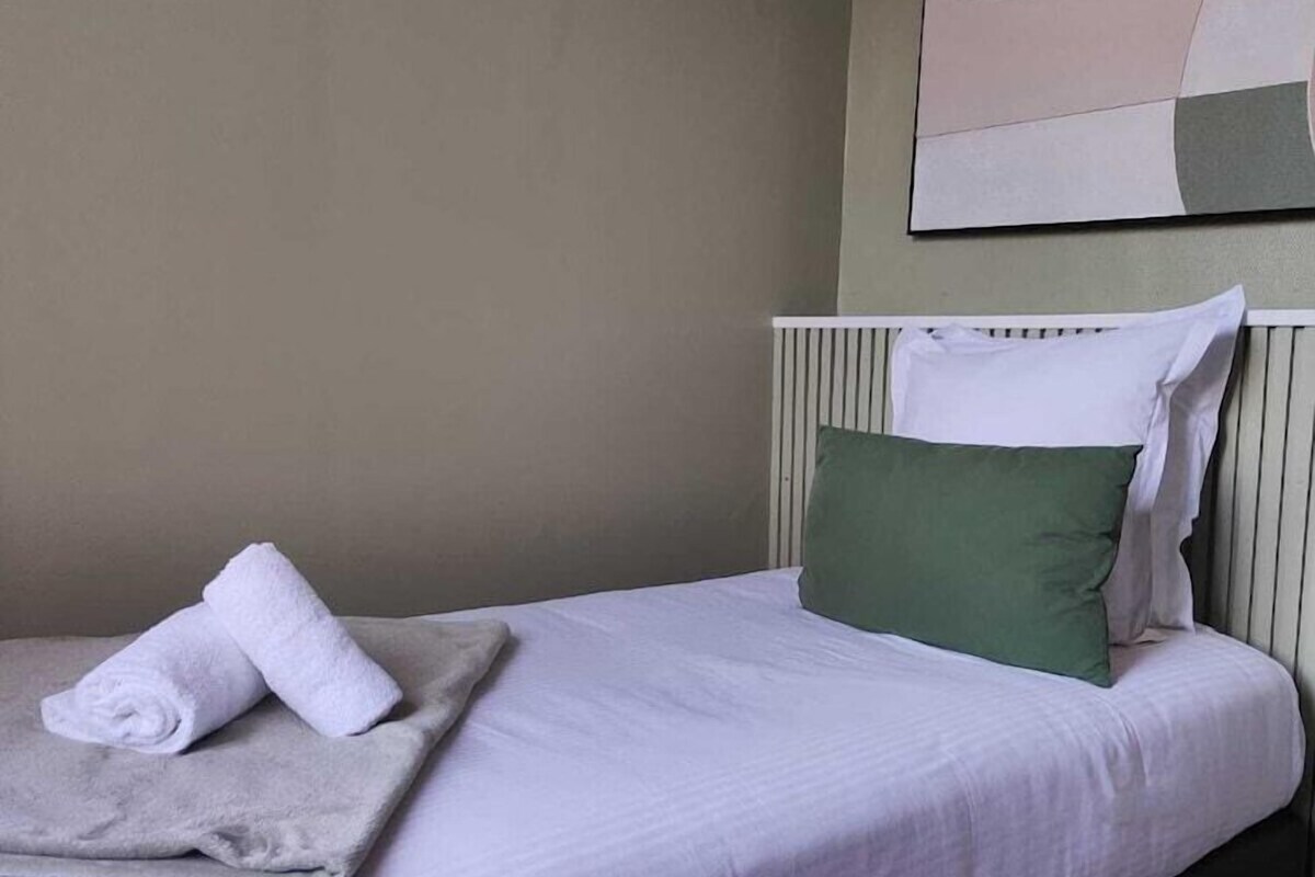 A neatly made bed with a white comforter and a green pillow, a rolled white towel on a blanket, and a wall with a simple abstract painting.