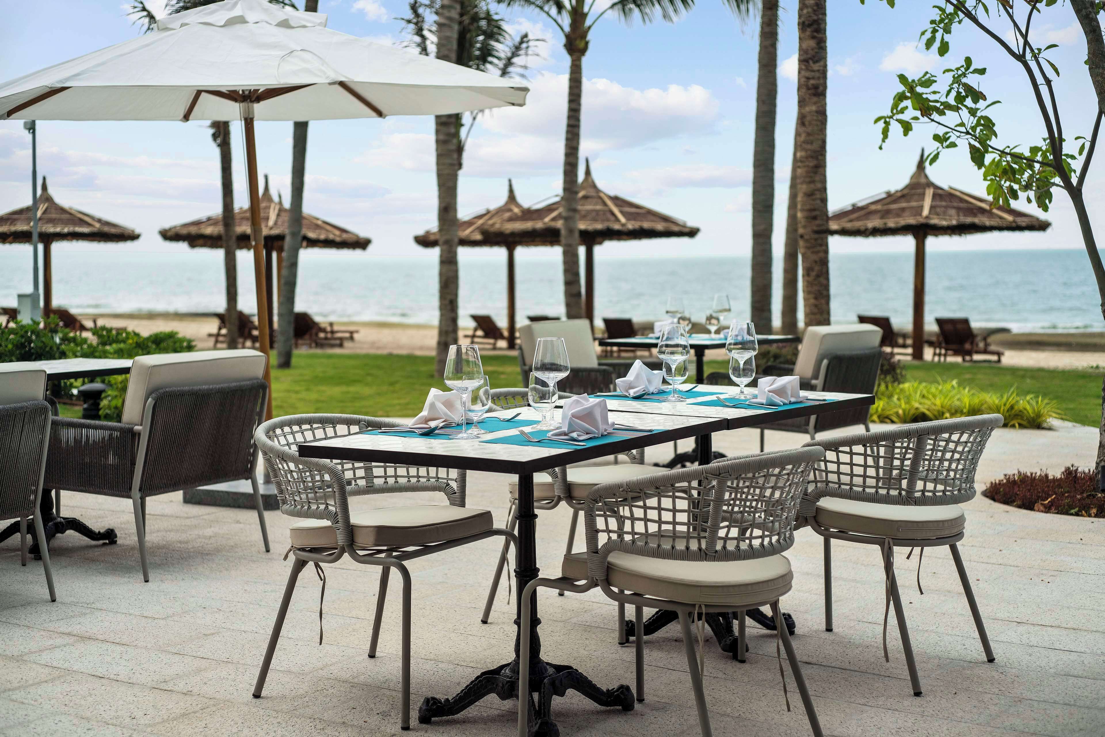 An outdoor dining area with a table set for four, surrounded by wicker chairs, with a view of the beach and ocean in the background.