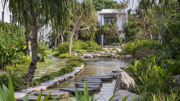 A garden with a stone bridge over a stream, surrounded by lush greenery and a modern building in the background. - Angsana Ho Tram (Ho Chi Minh City)