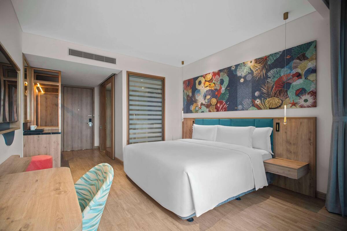 A modern hotel room with a large bed, a wooden headboard, a colorful abstract painting, a small table, and a bench.