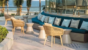 Un área de estar moderna al aire libre con sofá, sillas y mesas, con vistas a una playa y palmeras.