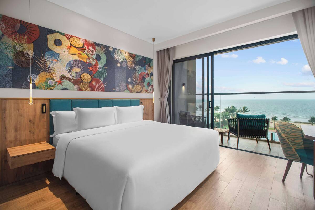 A hotel room with a large bed, a colorful mural, a balcony with a view of the sea, and a chair.