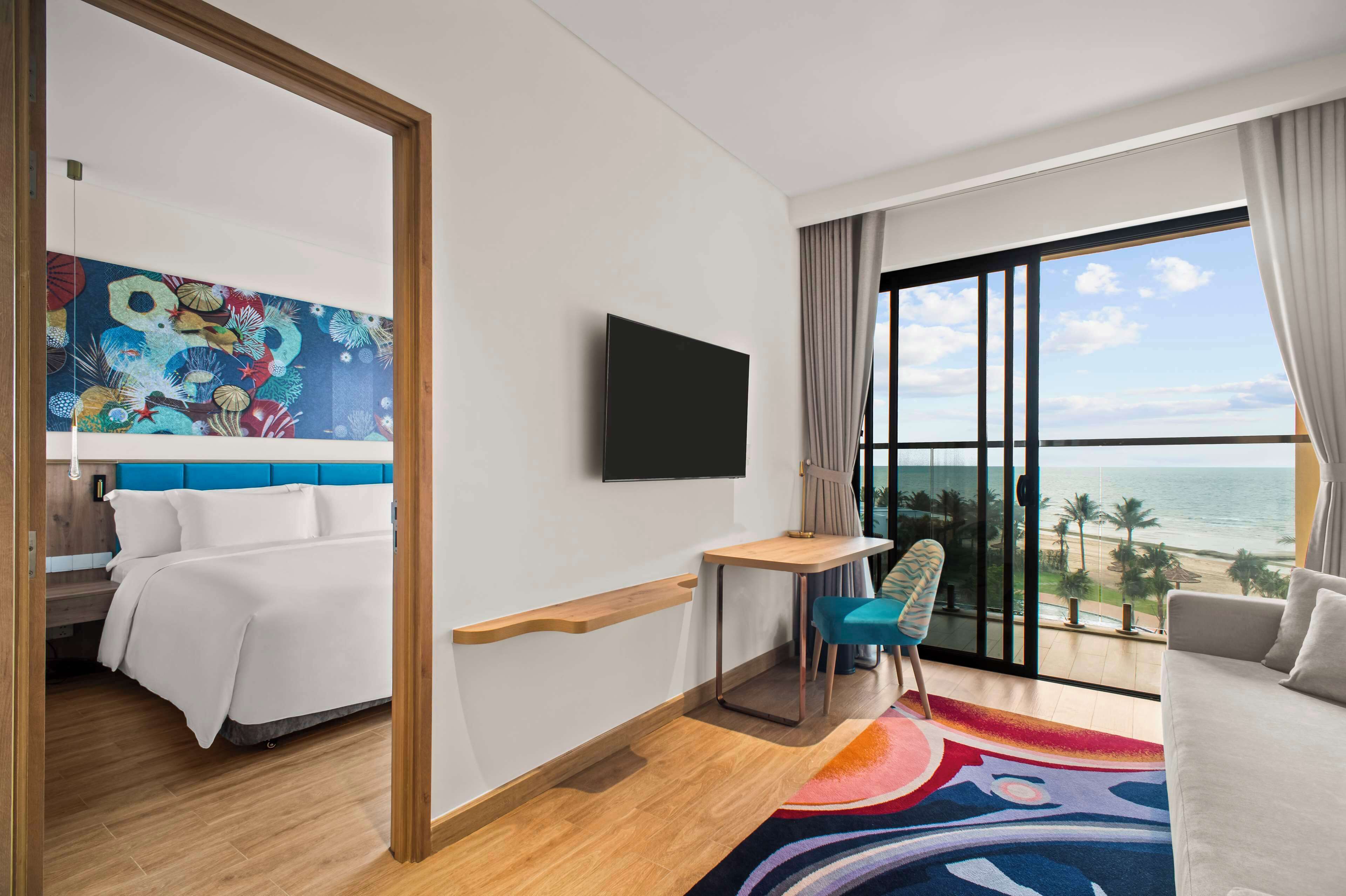 Habitación de hotel con cama, escritorio, silla, televisor y un amplio ventanal con vista a la playa.