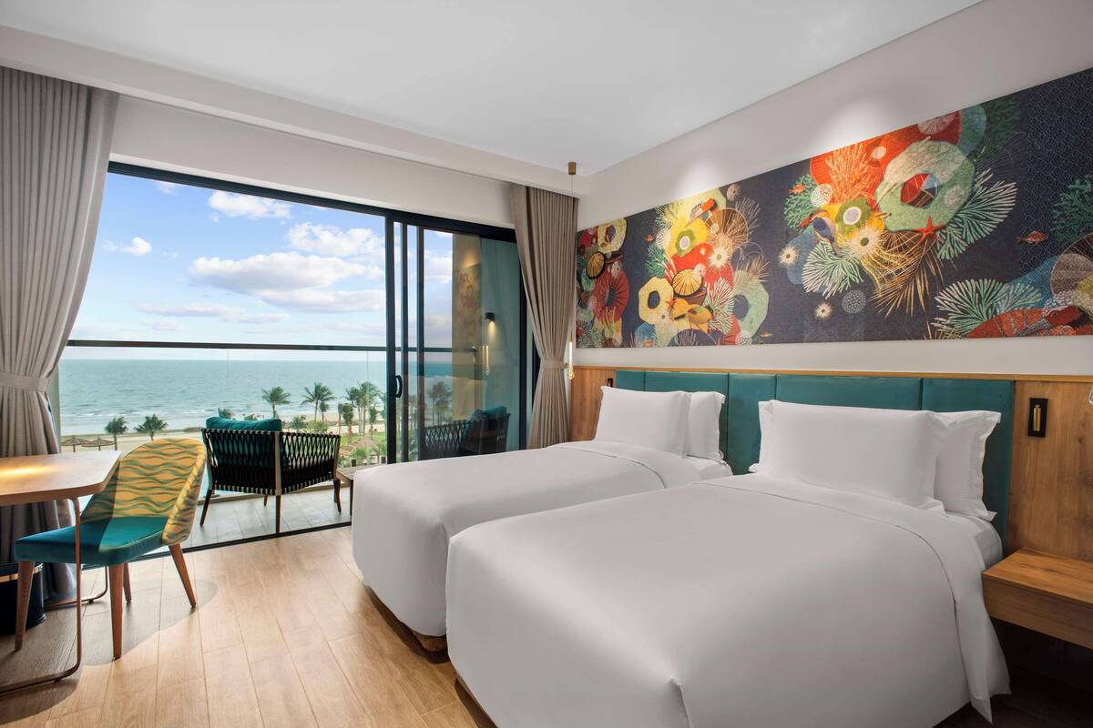 A hotel room with two beds, a dining area, and a balcony with a view of the ocean.