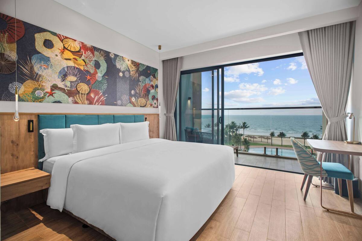 A hotel room with a large bed, a view of the ocean, a dining area, and a colorful mural on the wall.