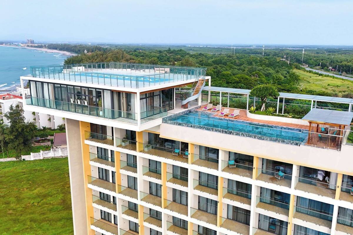 A modern hotel with a rooftop pool, multiple balconies, and a view of the coastline.