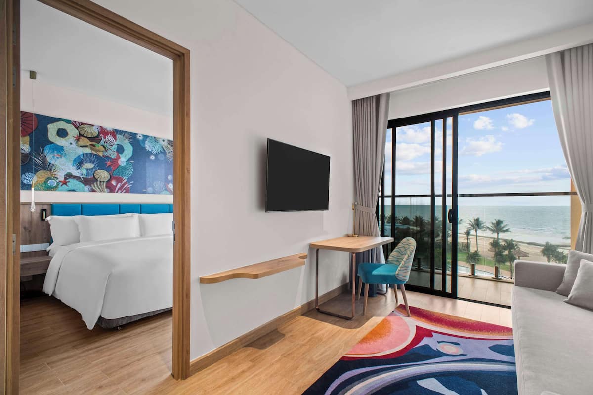 A hotel room with a bed, a desk, a chair, a TV, and a large window with a view of the beach.