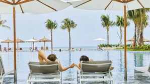 Un área junto a la piscina con sillones de descanso y sombrillas, palmeras y vista al océano.