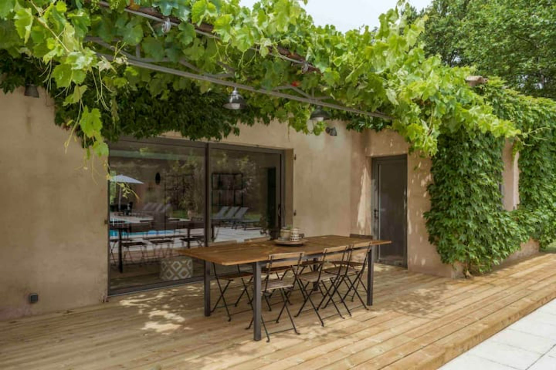 Une terrasse avec une table et des chaises en bois, une porte vitrée donnant sur une piscine et un mur recouvert de plantes grimpantes vertes.