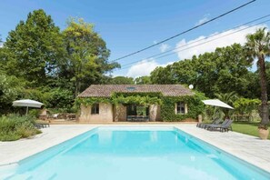 A swimming pool with a tiled deck, a small building with a tiled roof and ivy, and two umbrellas.
